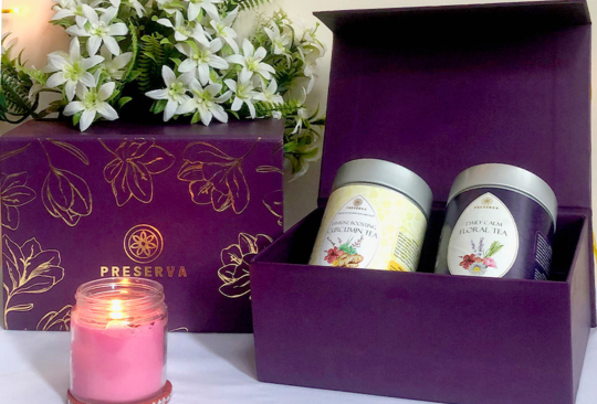 Two boxes of Preserva Wellness Tea gift boxes with two herbal teas next to white flowers and a pink candle