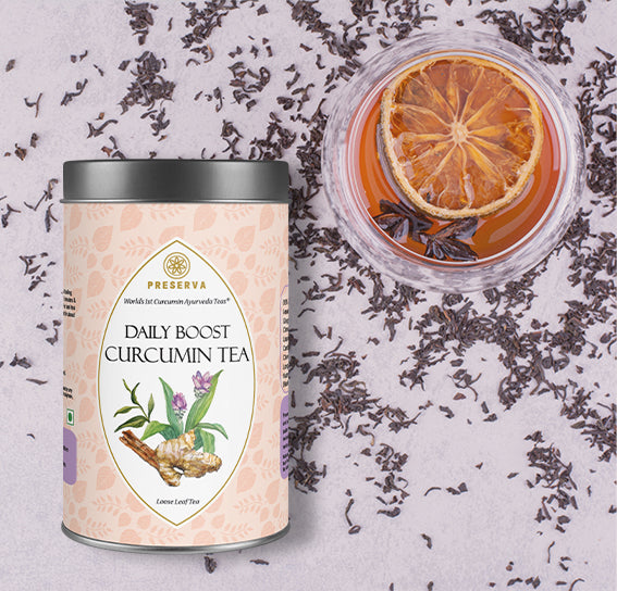 Preserva Wellness Daily Boost Curcumin Tea Box 50 grams next to a tea-filled glass cup with a slice of orange and star anise in it. The background has tea leaves spread out.