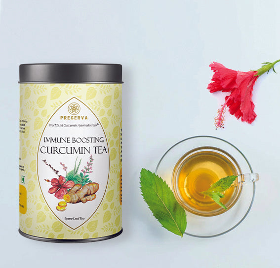 Immune Boosting Curcumin Tea Box 50 grams next to a glass cup with herbal tea with mint leaves and a hibiscus flower on a light blue background.